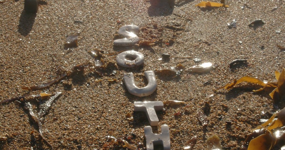 sand on a beach with stones, seaweed and silver letters spelling south